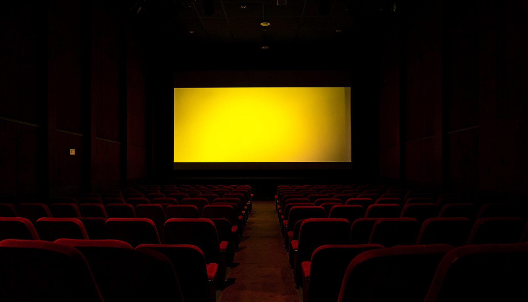 Darkened cinema auditorium with rows of red velvet seats, a glowing screen casting warm light across the room, deep shadows and rich crimson tones