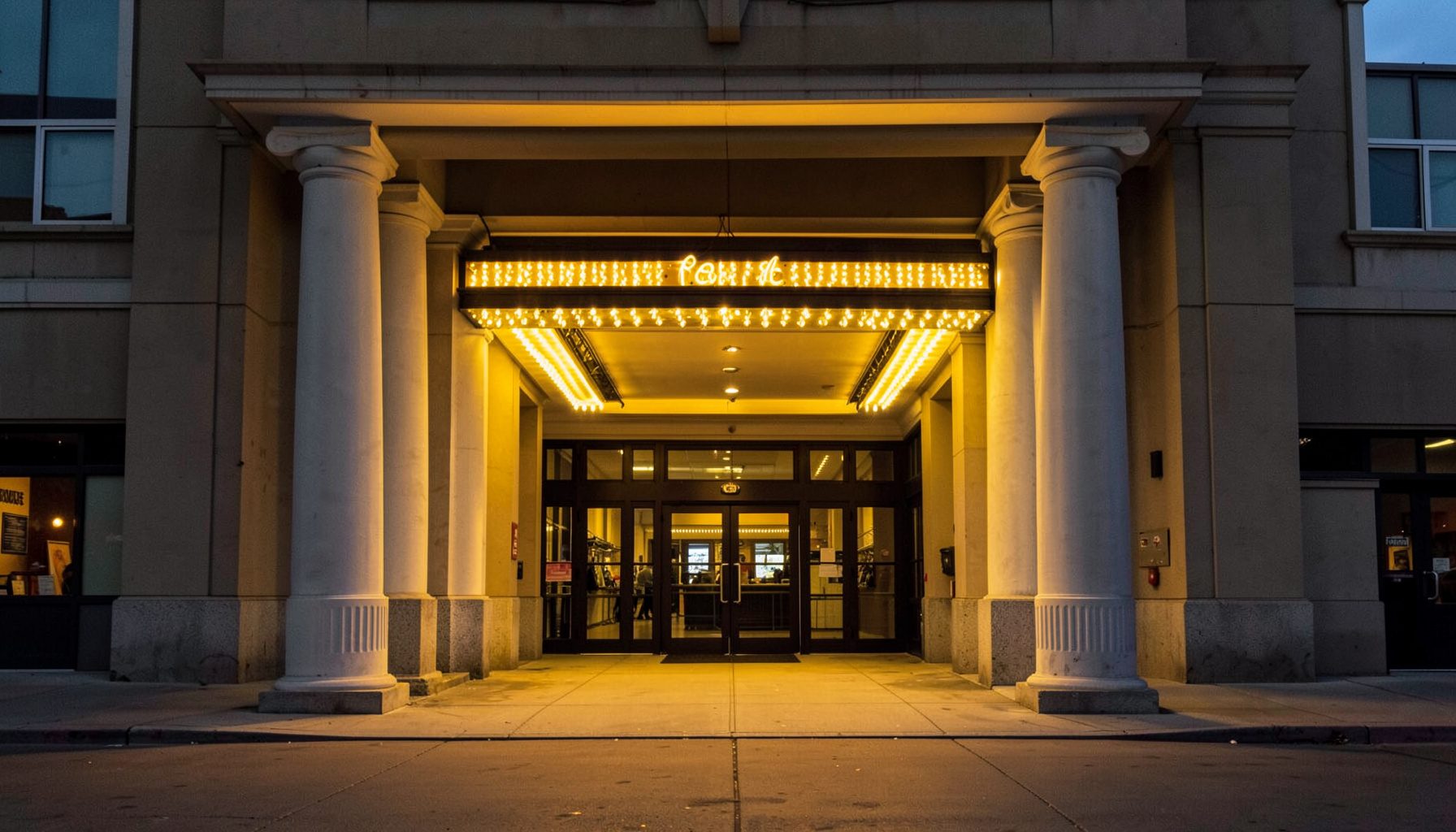 Grand movie theater entrance with symmetrical columns and an illuminated marquee, golden light spilling onto the sidewalk at dusk