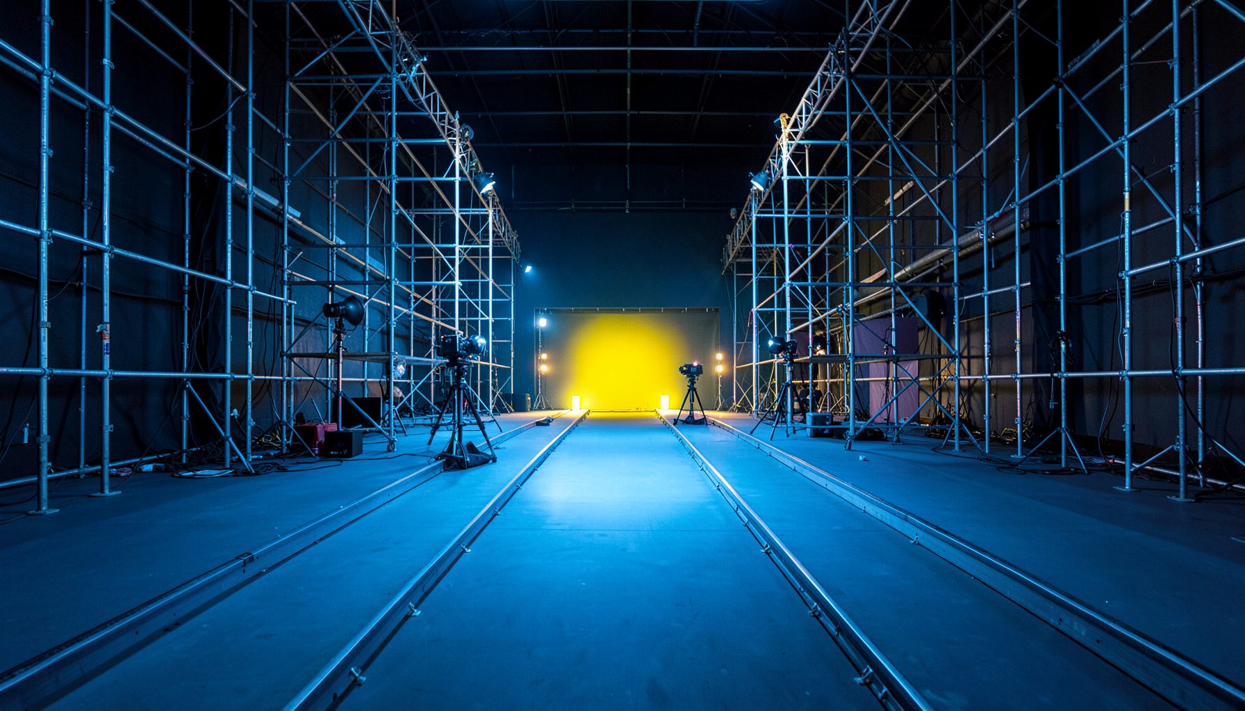 Wide-angle view of a film set with symmetrical scaffolding, lighting rigs, and a camera on a dolly track, bathed in cool tungsten blue and warm accent lights