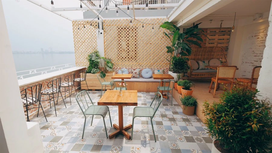 Rooftop terrace with patterned tile floor, wooden tables and chairs, potted plants and a hazy waterfront view, relaxed cafe atmosphere